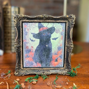 May include: A framed print featuring a black cat wearing a witch hat, surrounded by pumpkins and bats. The ornate, dark-toned frame adds a vintage aesthetic. A Halloween-themed decorative item.