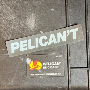 Könnte beinhalten: Ein dunkelgrauer Pelican 1510 Koffer mit einem Aufkleber mit der Aufschrift "PELICAN'T" in weißen Buchstaben. Ein kleinerer Aufkleber darunter lautet "PELICAN 1510 CASE" mit einem Logo und dem Text "PELICAN PRODUCTS, TORRANCE, CA USA".