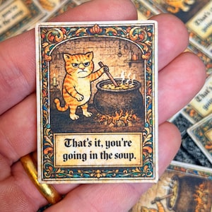 Puede incluir: Tarjeta rectangular con una ilustración de un gato atigrado naranja removiendo un caldero. La tarjeta tiene un borde adornado y el texto "That's it, you're going in the soup."