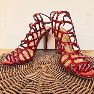 May include: A pair of red high-heeled sandals with a cage-like design. The open-toe shoes have a glossy finish and thin ankle straps. The heels are approximately 4 inches tall. The sandals are displayed on a woven surface.