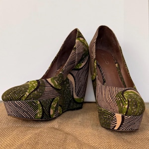 May include: A pair of wedge heels with a platform sole. The shoes feature a patterned fabric in shades of green, tan, and black. The heels are angled and the platform is thick. The shoes are displayed on a brown surface.