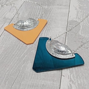 Puede incluir: Dos collares plateados con un colgante texturizado en forma de concha. Uno está sobre una base naranja en forma de diamante, el otro sobre una base verde azulado en forma de diamante. Ambos tienen cadenas plateadas.