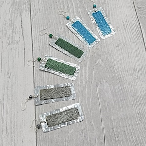 Puede incluir: Cinco pares de pendientes de plata con colgantes de forma rectangular. Los colgantes están hechos de metal y tienen una superficie texturizada. Los pendientes están decorados con piedras verdes y azules.