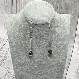 May include: A pair of silver dangle earrings with a long, thin silver bar and a small, round, dark red gemstone at the bottom.