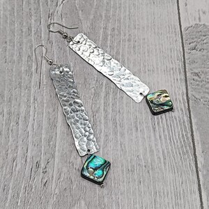 Puede incluir: Un par de pendientes de plata con colgantes de metal martillado y detalles de concha de abulón iridiscente.