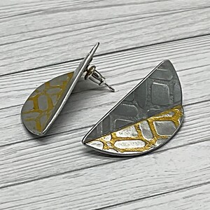 May include: A pair of silver and gold earrings with a geometric pattern. The earrings are shaped like half circles and have a textured surface.