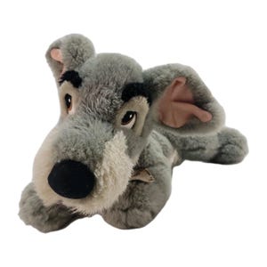 May include: A plush dog toy featuring soft gray fur, a cream-colored muzzle, and a black nose. The stuffed animal has large, expressive brown eyes, prominent black eyebrows, and floppy ears with pink inner lining, depicted in a lying position.