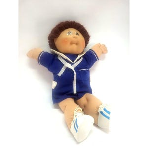 May include: A soft-sculpture doll with brown yarn hair, a round face, and rosy cheeks. The doll wears a dark blue collared shirt with white trim, a small pocket, and matching shorts. It is dressed in white sneakers with blue stripes on the sides.