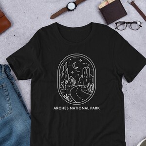 May include: Black t-shirt with a white outline of a desert landscape with a road, mountains, and a crescent moon. The text "ARCHES NATIONAL PARK" is printed below the image.