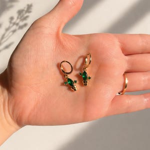 Puede incluir: Un par de pendientes de aro dorados con un pequeño diseño de cactus de piedra preciosa verde. Los pendientes se sostienen en una mano y se ve un anillo dorado en un dedo. Diseño sencillo y elegante.