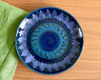 May include: A round, ceramic plate with a vibrant, concentric design in shades of blue and teal. The plate features a central dark blue circle surrounded by lighter blue and teal patterns, with a textured, star-like border. The plate is on a wooden surface.