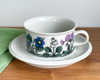 May include: A white teacup and saucer with a floral pattern. The pattern features blue, white, and purple flowers with green leaves.