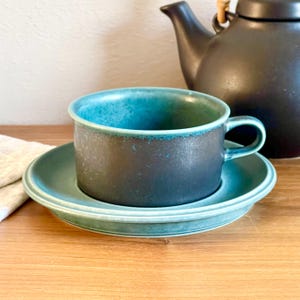Puede incluir: Una taza de té de cerámica en color verde azulado y gris oscuro con su plato a juego. La taza presenta una forma cilíndrica ancha con una sola asa. Una tetera negra mate se sitúa detrás del conjunto sobre una superficie de madera.