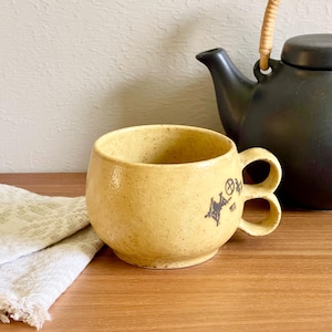 Könnte beinhalten: Ein gesprenkelter, hellgelber Keramikbecher mit rundem Körper und zwei übereinanderliegenden, kreisförmigen Henkeln. Der Becher hat ein kleines, dunkelbraunes Stempeldesign an der Seite. Dahinter steht eine mattschwarze Teekanne mit geflochtenem Henkel.