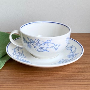 Op de afbeelding: Een wit theekopje en schotel met een blauw bloemenmotief. Het kopje heeft een ronde vorm en een handvat. De schotel heeft een gegolfde rand.