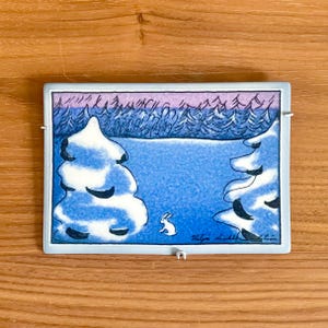 Op de afbeelding: Een rechthoekige keramische tegel met een winterse scène. Het kunstwerk toont twee met sneeuw bedekte dennenbomen die een besneeuwd landschap omlijsten met een klein wit konijn. De achtergrond toont een bos en een paarsblauwe lucht.