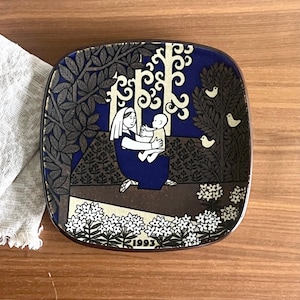 May include: Square ceramic plate with a dark blue background and a decorative illustration. The illustration depicts a woman in a blue dress holding a baby, surrounded by trees and flowers. The plate has a brown border and the year "1993" is printed on the bottom.