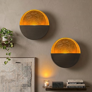 May include: Two wall sconces with a half-circle design. The top half is a warm gold color, and the bottom half is a dark bronze. The sconces are mounted on a neutral-colored wall. The image also shows a small shelf with a candle and books.