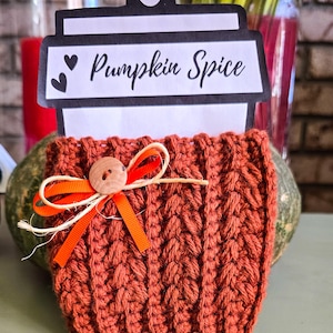 May include: A handmade, russet-coloured crochet coffee cup cosy with a wooden button and orange ribbon. The cosy holds a paper cup-shaped sign that reads "Pumpkin Spice" in black script. The cosy is decorated with a small orange bow.