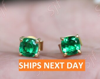May include: A pair of gold earrings with square emerald green gemstones. The earrings are on a hand.