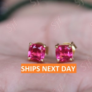 Könnte beinhalten: Ein Paar quadratische Ohrstecker mit leuchtend rosa Edelsteinen und goldfarbenen Fassungen. Die Ohrringe werden vor einem verschwommenen Hintergrund präsentiert. Der Text "SHIPS NEXT DAY" ist in einer orangefarbenen Box zu sehen.