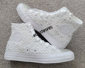May include: White high-top sneakers covered in white pearls and rhinestones. The sneakers have white satin laces and a white star logo on the side.