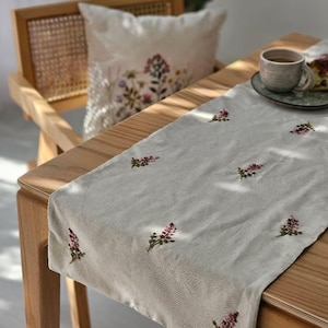 May include: A light beige linen table runner with embroidered sprigs of purple flowers and green leaves. A matching pillow with floral embroidery rests on a wooden chair. A cup of coffee and a small plate are on the table.