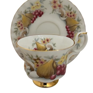 Puede incluir: Un juego de taza y platillo de té vintage. La taza y el platillo de porcelana blanca tienen un borde dorado y están decorados con un patrón de frutas y flores en tonos amarillos, marrones y rojos. La taza tiene una base y un asa doradas.