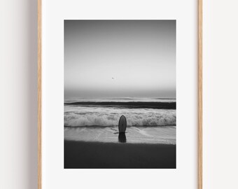 May include: Black and white framed photograph of a surfboard on a sandy beach at dusk. The ocean waves are crashing in the background under a cloudy sky. A bird flies in the distance.