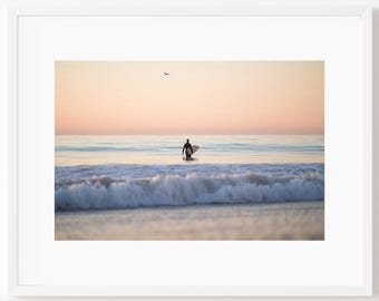 May include: A framed photograph of a surfer walking into the ocean at sunset. The sky transitions from pink to yellow, with a bird flying overhead. The surfer is silhouetted against the water, holding a surfboard.