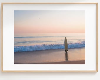 May include: A framed print of a beach scene at sunset. The sky transitions from pink to blue over the ocean. A surfboard stands upright in the sand near the water's edge. The frame is a light wood color.