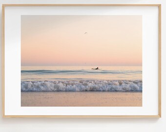 May include: A framed print of a beach scene. The image features a surfer in the ocean, with waves breaking on the shore. The sky is a soft pink and orange, and a bird flies overhead. The frame is a light wood color.