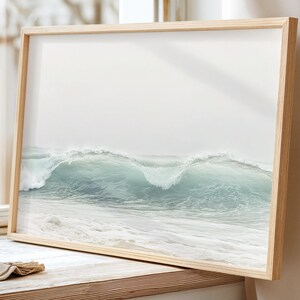 May include: A framed print featuring a serene ocean wave in shades of teal and white. The artwork is displayed in a light wood frame, capturing the essence of coastal decor. The image is set against a window with natural light.