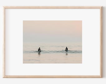 May include: A framed print featuring a serene ocean scene. Two figures are silhouetted in the water, likely surfing or waiting for waves. The sky has a soft, pastel color palette, creating a calming and minimalist aesthetic.