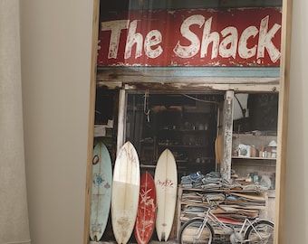 Könnte beinhalten: Gerahmter Druck eines Surfshops mit dem Text "The Shack" in Rot und Weiß. Mehrere Surfbretter lehnen an dem Gebäude. Ein Fahrrad steht in der Nähe. Das Bild hat eine Vintage- und Strandatmosphäre.