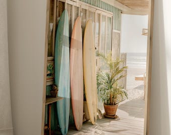 May include: A framed print featuring three surfboards in pastel colors: light blue, pink, and yellow. The surfboards lean against a weathered wooden structure with a beach scene visible in the background. A potted plant sits nearby.