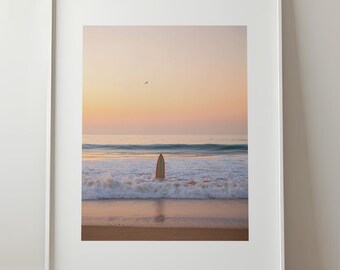 May include: A framed print of a surfboard standing in the ocean at sunset. The surfboard is vertical in the water, with waves crashing around it. The sky is a gradient of orange and pink, and the sand is a light brown.