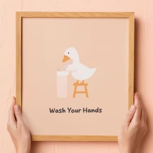 May include: A framed print featuring a cartoon duck washing its hands. The duck is white with an orange beak, standing on a small stool. The print has the text "Wash Your Hands" in black font on a peach background.