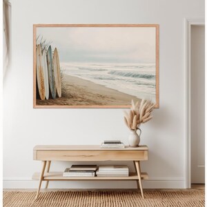 May include: A framed print depicts a serene beach scene with surfboards leaning against the dunes. The artwork features a sandy beach, ocean waves, and a muted color palette. A wooden console table with books and a vase of pampas grass sits below the artwork.