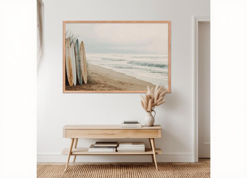 May include: A framed print depicts a serene beach scene with surfboards leaning against the dunes. The artwork features a sandy beach, ocean waves, and a muted color palette. A wooden console table with books and a vase of pampas grass sits below the artwork.