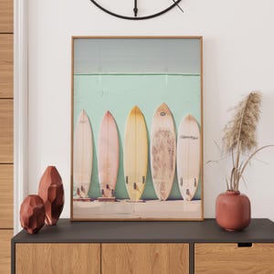 May include: A framed print featuring five vintage surfboards leaning against a teal wall. The surfboards are various pastel shades of yellow, pink, and orange.  The image evokes a beachy, summer vibe, ideal for home decor or a beach-themed business. The surfboards are arranged neatly, suggesting a calm and organized aesthetic. The print is a great addition to any space needing a touch of coastal charm.