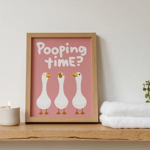 May include: A framed print with the text "Pooping time?" above three cartoon geese on a pink background. A lit candle and rolled white towels sit on a wooden shelf.