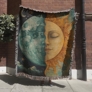 May include: A woven tapestry blanket featuring a celestial design. One half depicts a serene sun with closed eyes, the other a moon with stars and clouds. The blanket has a fringed edge and is primarily teal, gold, and navy blue.