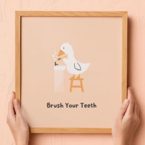 May include: A framed print featuring a cartoon duck brushing its teeth. The duck is white with an orange beak and stands on a small stool. The text "Brush Your Teeth" is below the illustration. The frame is made of light brown wood.