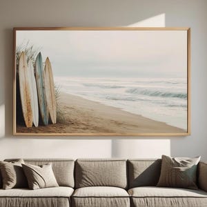 May include: A framed print featuring a beach scene with three surfboards leaning against dune grass. The surfboards are weathered with shades of brown, blue, and white. The ocean waves and sandy beach are visible in the background, creating a coastal theme.