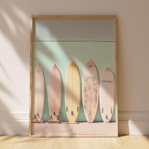 May include: A framed print featuring five vintage surfboards leaning against a pale green wall. The surfboards are in various pastel shades of yellow, beige, and light brown.  The image evokes a beachy, summery vibe, ideal for coastal-themed home decor. The surfboards are displayed in a row, creating a visually appealing composition. The print is perfect for adding a touch of retro style to any room.