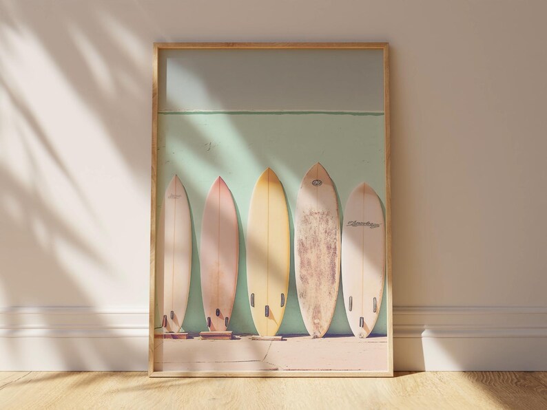 May include: A framed print featuring five vintage surfboards leaning against a pale green wall. The surfboards are in various pastel shades of yellow, beige, and light brown.  The image evokes a beachy, summery vibe, ideal for coastal-themed home decor. The surfboards are displayed in a row, creating a visually appealing composition. The print is perfect for adding a touch of retro style to any room.