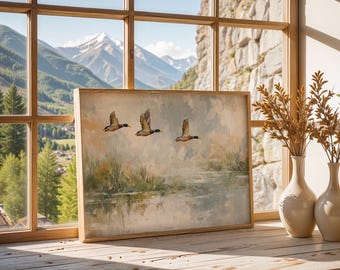 May include: A framed landscape painting depicts three ducks in flight over a body of water, with a backdrop of marshy vegetation and a soft, cloudy sky. The artwork is set against a window with a mountain view, and is displayed on a wooden surface.