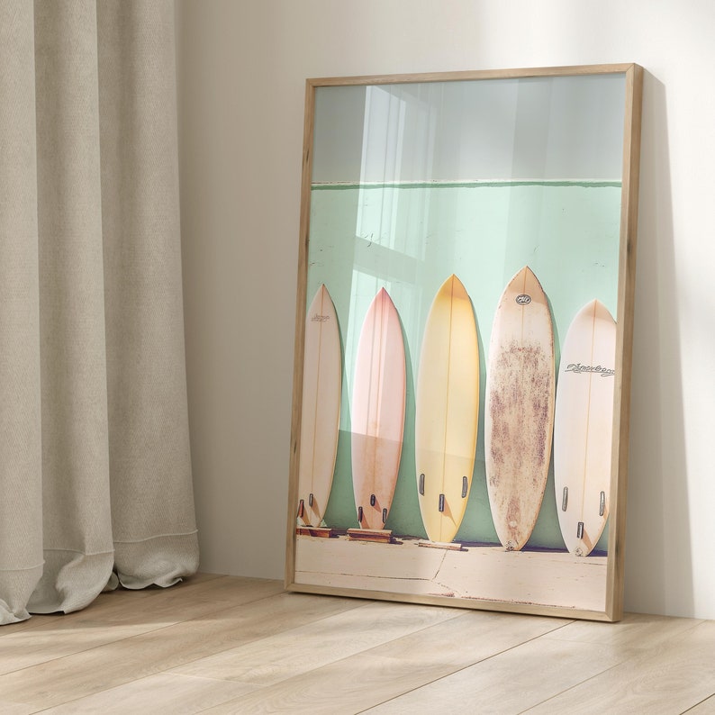 May include: A framed wall art print featuring six vintage surfboards in various pastel colors, including yellow, pink, and beige, lined up against a light teal wall. The surfboards have a worn, vintage look, and some have visible text and branding. The overall style is reminiscent of beach culture and summer vibes. The frame is light wood, and the image is well-lit, creating a bright and airy feel.