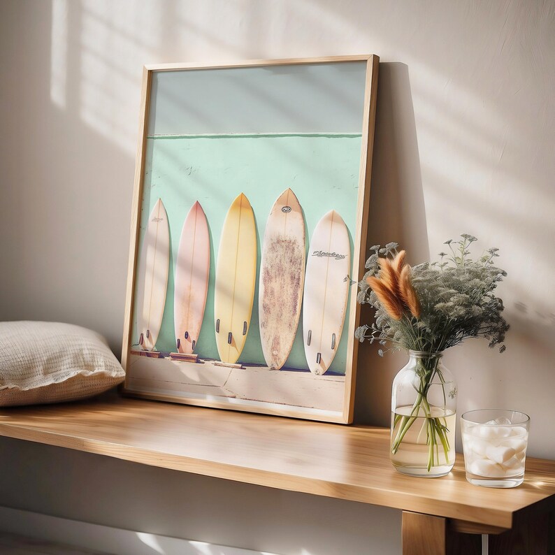 May include: A framed print featuring five vintage surfboards leaning against a light teal wall. The surfboards are various shades of yellow, beige, and light brown. The print is in a light wood frame and sits on a light wood surface.  The overall style is beachy and evokes a sense of summer.